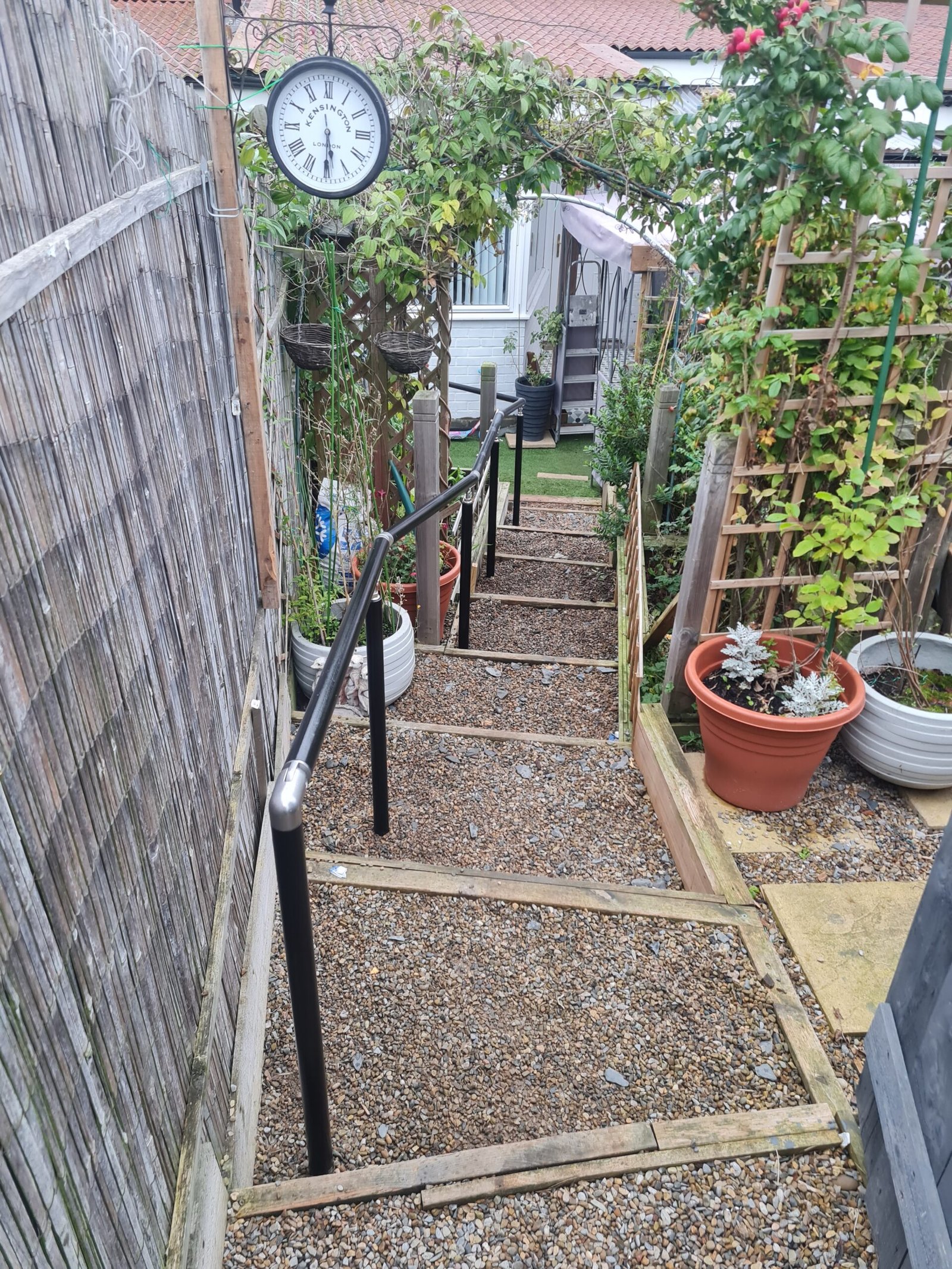 Garden steps leading down with a handrail, surrounded by plants and a clock on the wall, showcasing outdoor landscaping features.